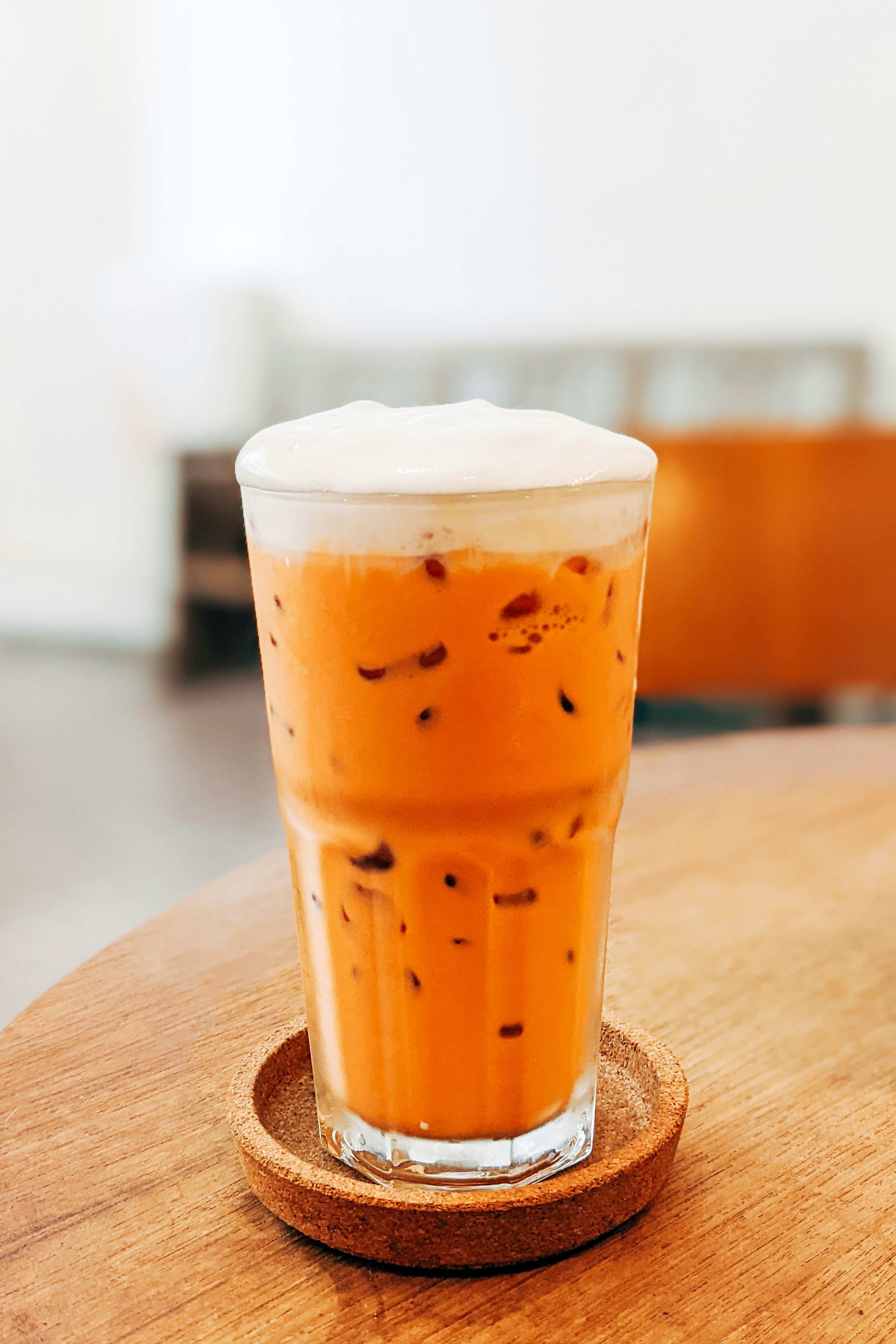 A glass of Thai tea with foam on top sits on a coaster on a wooden table.